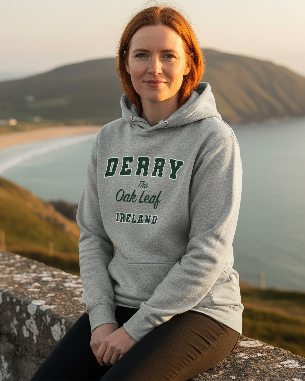 Person wearing a gray hoodie with 'Derry The Oak Leaf Ireland' text, sitting outdoors with a scenic background.
